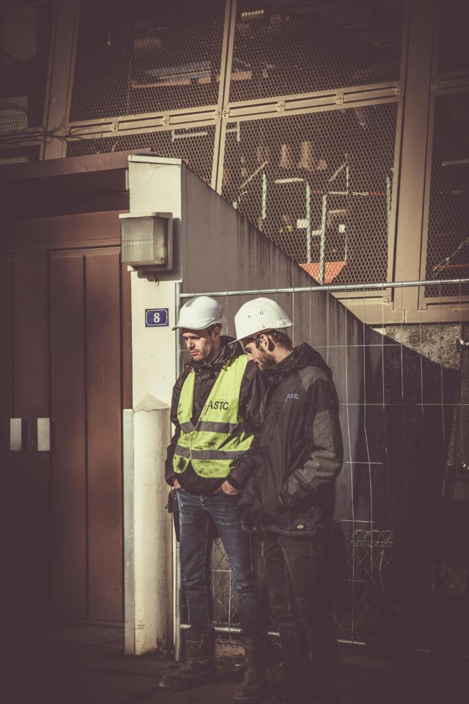 Two construction workers in safety gear standing outdoors at a building site, focused on work.